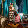 A child dressed in a Meri Meri Puppy Costume - Final Sale stands on a stage, holding a treat and pointing at a brown dog adorned in a pink frilly dress and party hat. Surrounding them are a drum, a tuba, and a disco ball, with blue curtains and balloon decorations in the background.