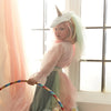 In a sunlit room, a young child stands by the window dressed in the Meri Meri Rainbow Unicorn Costume - Final Sale. The whimsical outfit features a velvet unicorn headpiece adorned with a shining horn and is paired with a colorful pastel skirt and a vibrant rainbow tulle tail. Holding a multicolored hula hoop, the child gazes back at the camera as soft curtains frame the charming scene.