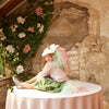 A young girl sporting the Meri Meri Rainbow Unicorn Costume - Final Sale, complete with a velvet unicorn headpiece and a rainbow tulle tail, sits on a round table covered with a pink tablecloth. She is surrounded by rustic decor, featuring flowers and ivy adorning a textured, worn wall. Green and pink tones prevail in the whimsical scene.