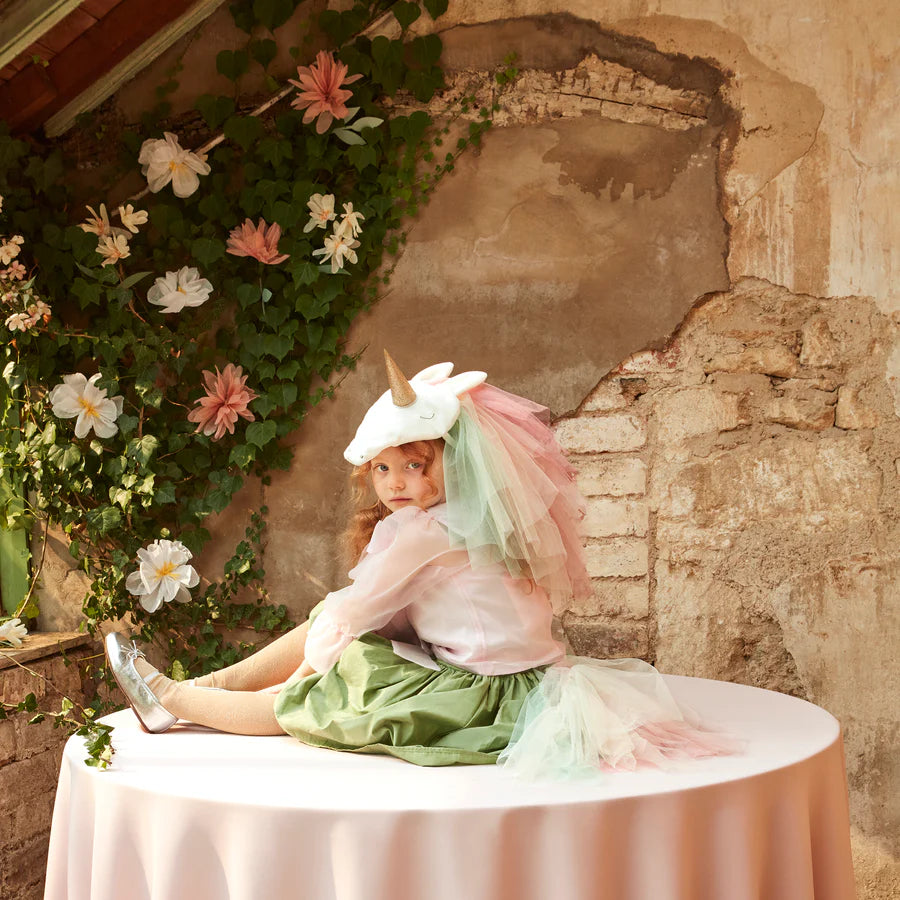 A young girl sporting the Meri Meri Rainbow Unicorn Costume - Final Sale, complete with a velvet unicorn headpiece and a rainbow tulle tail, sits on a round table covered with a pink tablecloth. She is surrounded by rustic decor, featuring flowers and ivy adorning a textured, worn wall. Green and pink tones prevail in the whimsical scene.