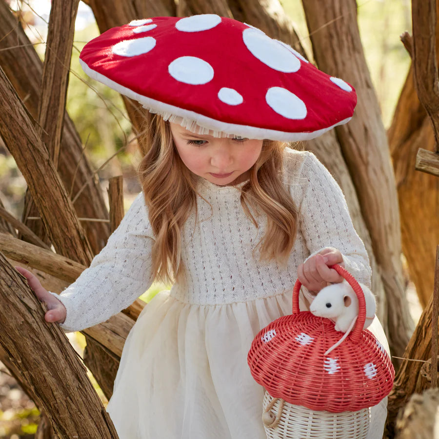 A young girl wearing a white dress and the Meri Meri Mushroom Hat, shaped like a red mushroom with white spots, is standing among tree branches. She is holding a small wicker basket, also designed as a mushroom, with a stuffed white mouse peeking out of it, creating an atmosphere of woodland wonderfully imaginative play.