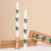 The MeriMeri Holly Table Candles, a set of two, feature a festive design with white wax adorned by green holly leaves and red berries. One candle is elegantly displayed in a brass holder, while its companion leans gently against a stack of brown boxes. A beige background enhances the classic charm and warmth of these traditional Christmas decorations.