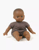 A brown-skinned baby doll with dark hair is seated on a white background. The Minikane Soft Body Doll (11") - Oscar, crafted from phthalate-free vinyl, is wearing a simple brown romper and has an expressive face with big dark eyes. Its legs are spread apart and arms are slightly open, perfect as a bedtime companion.