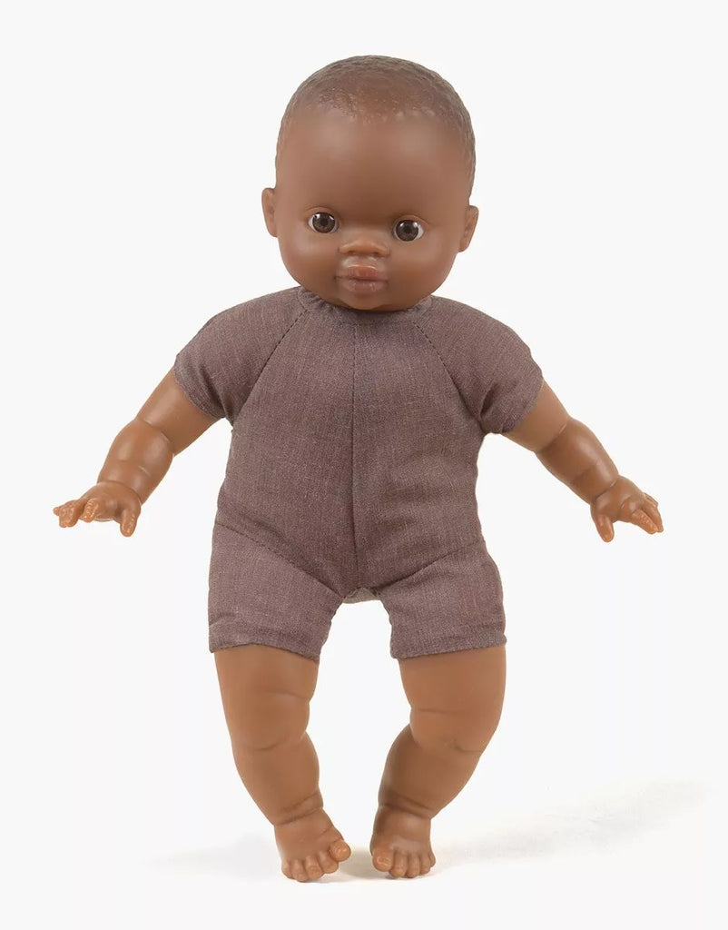 A Minikane Soft Body Doll (11") - Oscar with dark skin stands upright in a brown romper, arms slightly bent and legs positioned as if taking a step. Wide-open eyes and a neutral expression add realism. Made from phthalate-free vinyl, this doll looks perfect against the plain white background.