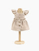 A wicker Minikane Mannequin For Doll Clothing displays a beige baby dress with ruffled sleeves and wooden buttons. The dress, designed for Minikane Gordis dolls, features a gathered waist and is made of a light, airy fabric—perfect for any stylish dressing room display.