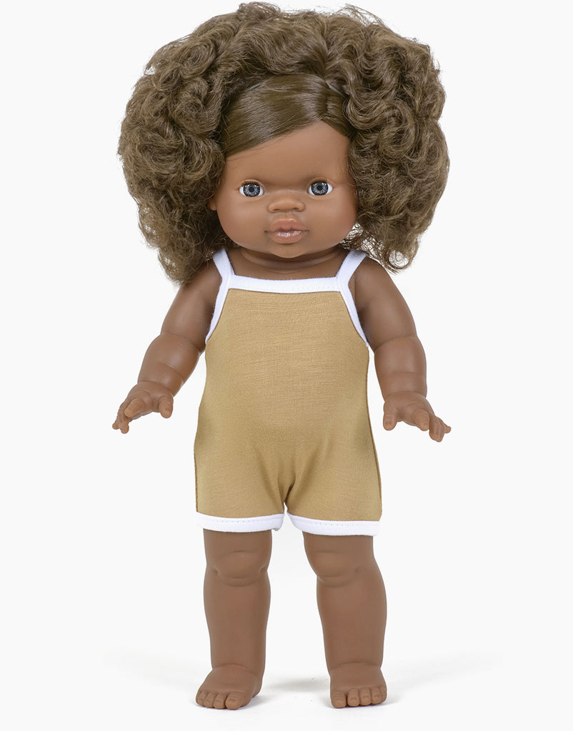 A Minikane Clothed Standing Doll (14.5") - Charlie with dark brown curly hair and brown skin stands against a white background. The doll, anatomically correct and with a gentle vanilla scent, is wearing a tan romper with white trim. Its eyes are open wide, and its arms are slightly extended outwards.