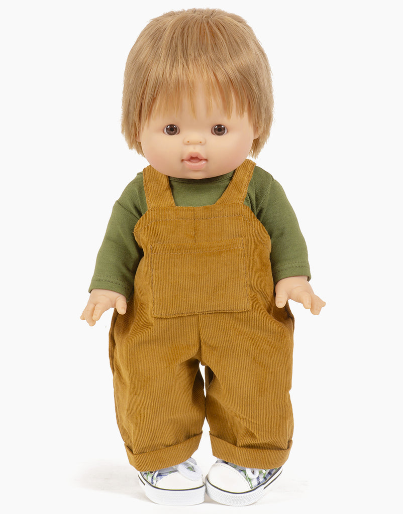 A Minikane Gordis doll with brown hair stands facing forward, dressed in an olive green jersey t-shirt and the Antoine Corduroy Overalls Set, which includes mustard yellow corduroy overalls. The doll also wears white shoes with a plaid pattern.