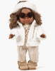 The Minikane Doll Clothing | Natalie Set features a doll dressed in a charming ensemble consisting of a white fluffy jacket, beige pants, and brown boots. Adorned with curly dark hair, round sunglasses, and a white headband, this exquisite piece is designed in France. Its knit composition enhances its appeal as it stands elegantly against a plain background.