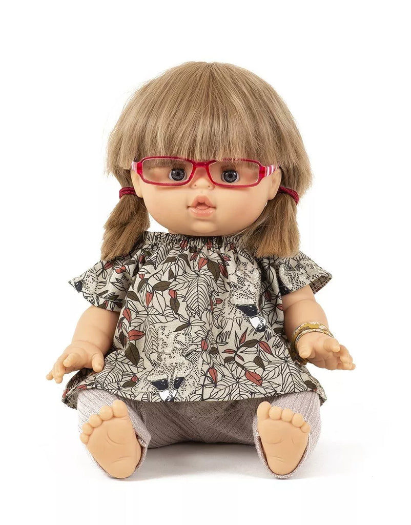 A doll with light brown hair tied in pigtails, wearing TESSA Doll Glasses by Minikane, a floral patterned dress, and light-colored leggings. The doll is seated with legs crossed and has an expressive face, perfect for imaginative play while prioritizing children's safety.