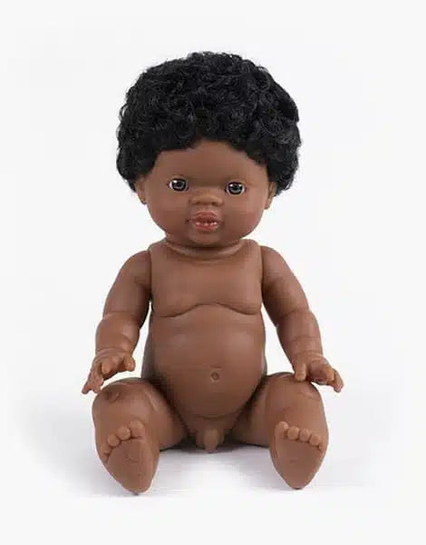 A brown-skinned, anatomically correct Minikane Baby Doll (13") - Jaro with curly black hair sits upright against a white background. The doll has a smiling expression and detailed facial features. It is unclothed and has its legs slightly apart and arms relaxed at its sides.