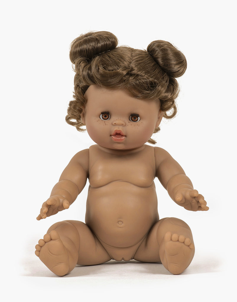 A brown-skinned Minikane Baby Doll With Sleeping Eyes (13") - Madeleine with curly brown hair styled in two buns sits with legs spread and arms reaching forward. The anatomically correct doll has detailed facial features, including brown eyes, small lips, and subtle freckles, and is undressed.