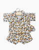 A child's two-piece clothing set on a hanger. The outfit includes a short-sleeved button-up shirt and matching shorts, both featuring a lively print of orange fruit and green leaves on a white background, perfect for pairing with Minikane Doll Clothing | Pajamas and Eye Mask in Orange Blossom.