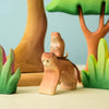 The Bumbu Wooden Monkey Set features handmade monkey figurines depicted in a forest-like setting with stylized trees and foliage. One monkey is portrayed riding on the back of another. Designed as eco-friendly toys, both the figures and background elements are painted in soft, earthy colors, creating a whimsical and playful scene.