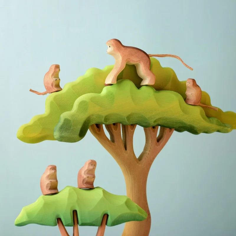 The Bumbu Wooden Monkey Set is a whimsical collection featuring two wooden trees with leafy canopies. Four handmade monkeys are playfully positioned on and around the trees, with one monkey perched on top, another standing, and two sitting on the branches. This eco-friendly toy set is presented against a light blue background.