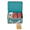 A teal and white suitcase is open, revealing two pink cups, a cloth with floral embroidered details, and a pair of novelty sunglasses. The suitcase has a latch and handle, with a tag that reads “Dinkum Doll - Sprout (Extended Pack).”