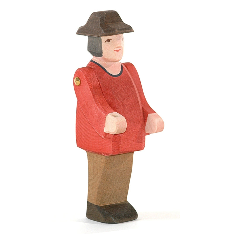 A wooden figurine of a person wearing a red shirt, brown pants, and a brown hat. The figure has simple features, articulated arms, and a flat base, enabling it to stand upright. Perfect for imaginative play with handcrafted wooden toys from the Ostheimer brand. Introducing the Ostheimer Farmer.