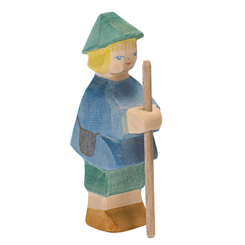 The Ostheimer Shepherd Boy is a handcrafted wooden figurine, likely representing a shepherd. The figure is painted in blue and green with a square hat, blond hair, and carries a wooden staff. Made from sustainably sourced materials, this piece features simple facial details with red lips and blue eyes.