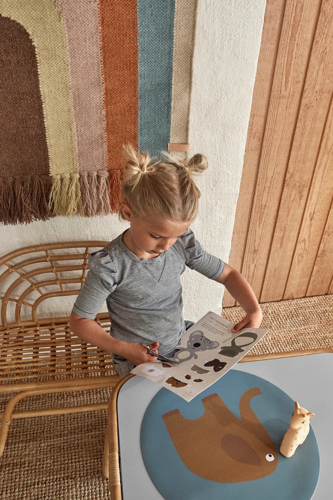 A young child with blonde hair tied in pigtails is standing by a small table, cutting out animal shapes from a piece of paper with scissors. An elephant-shaped placemat and a toy giraffe sit on the table. The background features rattan furniture, including a Kid’s Rattan Bench and a colorful woven wall hanging.