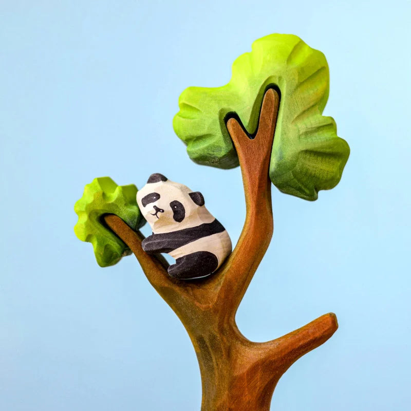 A Bumbu Panda Bears And Tree Set sits in the branches of a wooden tree with two green leaves. The background is a light blue. The panda has black and white markings, typical of its species, and is nestled on a branch, looking content—perfect for imaginative play scenes.