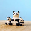 A Bumbu Panda Bears And Tree Set: an adult sits in the middle with two smaller pandas, one each on its left and right. All have black and white painting and expressive faces. The eco-friendly wood figurines rest on a light wood surface against a blue background.