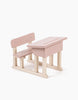 A small, powder pink wooden Minikane Doll School Desk with an attached bench. This set features a minimalistic design with unpainted wooden legs and braces, reminiscent of items crafted for Minikane Babies, and is set against a plain white background.