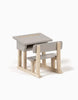 The Minikane Doll School Desk - Grey is a small, gray and light wood two-seater desk for children, reminiscent of a classic wooden school desk. The sloped top surface features a slot for storage underneath. Crafted with care by an artisan carpenter, it includes an attached bench-style seat for a compact and functional design. The background is plain white.