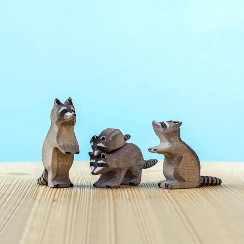 Three wooden raccoon figurines are placed on a table against a light blue background. This Bumbu Wooden Raccoon Set includes one raccoon standing on its hind legs, two stacked in the middle, and one sitting upright. Perfect for imaginative play, each piece is expertly handcrafted.