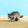 A figurine from the Bumbu Wooden Raccoon Set stands on a wooden surface against a light blue background. As part of an imaginative play handcrafted set, it features intricate details with black and white markings characteristic of a raccoon.