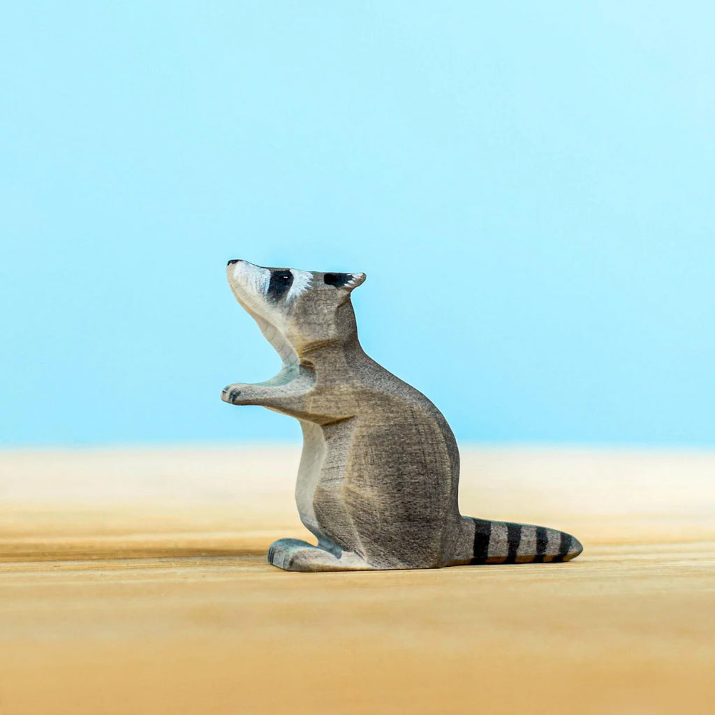 The Bumbu Wooden Raccoon Set includes a charming, small carved wooden raccoon standing on its hind legs against a backdrop that transitions from a light blue sky to a sandy beige foreground. Perfect for imaginative play, this handcrafted raccoon boasts distinct black and white markings on its face and a striped tail.