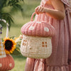 A person holding an Olli Ella Mushroom Basket - Pink, with a blurred background featuring greenery and a sunflower. The person is wearing a pink, ruffled dress.