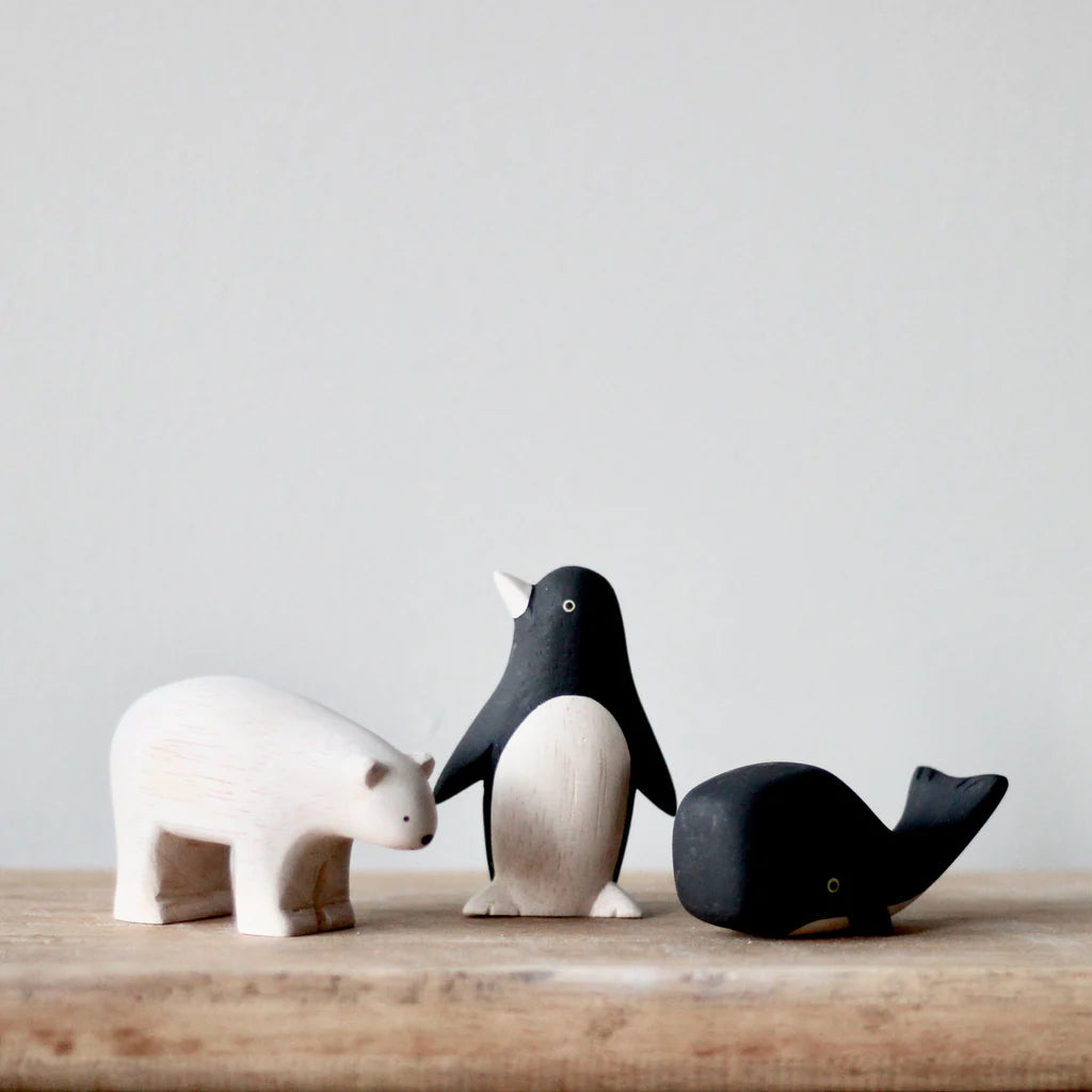 A Handmade Tiny Wooden Penguin, white and black in color, is beautifully arranged on a wooden surface alongside a polar bear and a whale. The polar bear is white, and the whale is black. Against a plain light gray wall, these handmade wooden toys bring charm to any space.