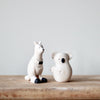 Two small, white ceramic figurines on a wooden surface against a plain background. One figurine is a kangaroo sitting upright with a black bow tie and black-tipped feet, while the other is the Handmade Wooden Koala with minimal facial features and short limbs, reminiscent of handcrafted wooden animals.