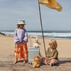 Two children dressed in casual outfits are at a beach with an Olli Ella See-Ya Suitcase - Steel Blue and toys, a yellow flag waving on a pole next to them, with the sea in the background.