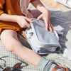 A person sitting on a sandy surface packing or unpacking a gray Olli Ella See Ya Wash Bag in Steel Blue, with a water bottle and sunglasses nearby, wearing a brown dress and white shoes.