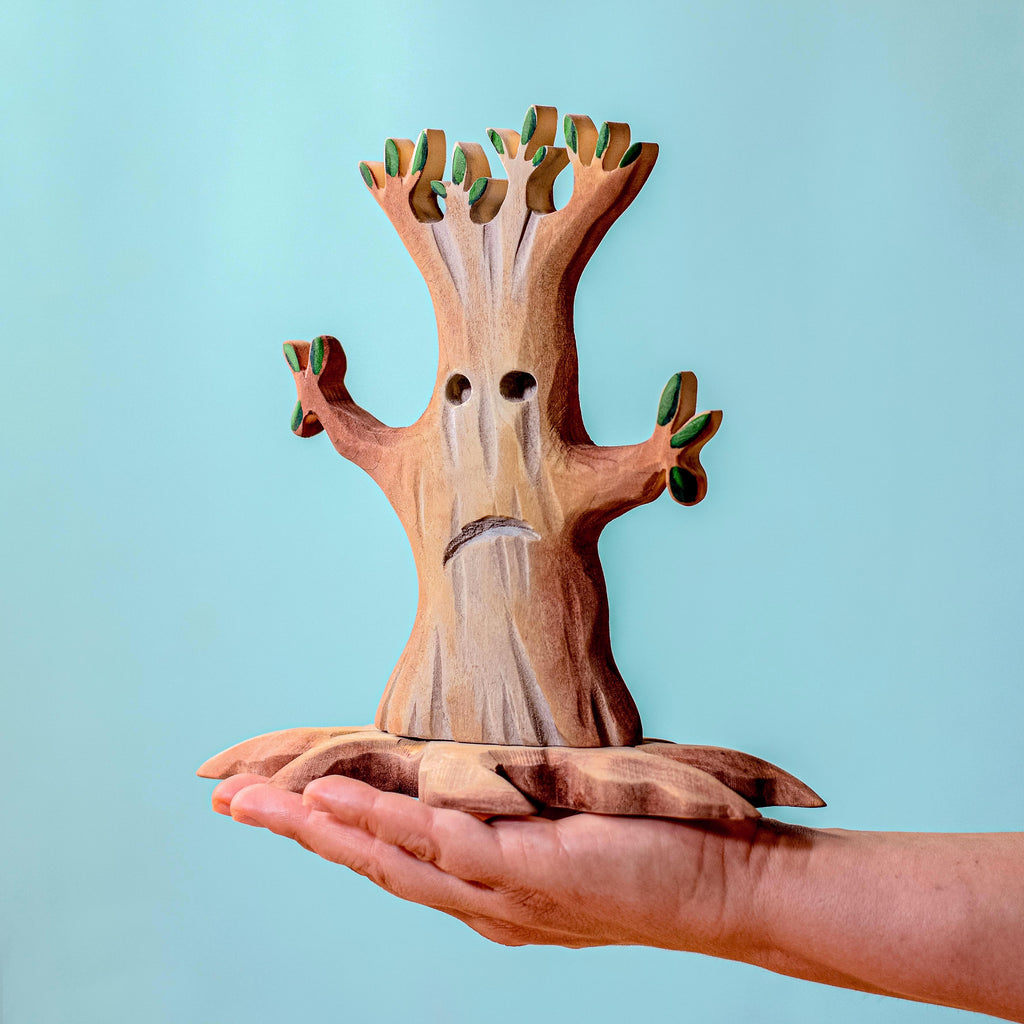 A person holds up a figure from the Handmade Wooden Halloween Trees & Bat Set against a solid light blue background. This handcrafted wooden tree, with its sad face, large expressive eyes, and drooping branch-like arms accented by green leaves and a detailed base, makes for perfect Halloween decor.