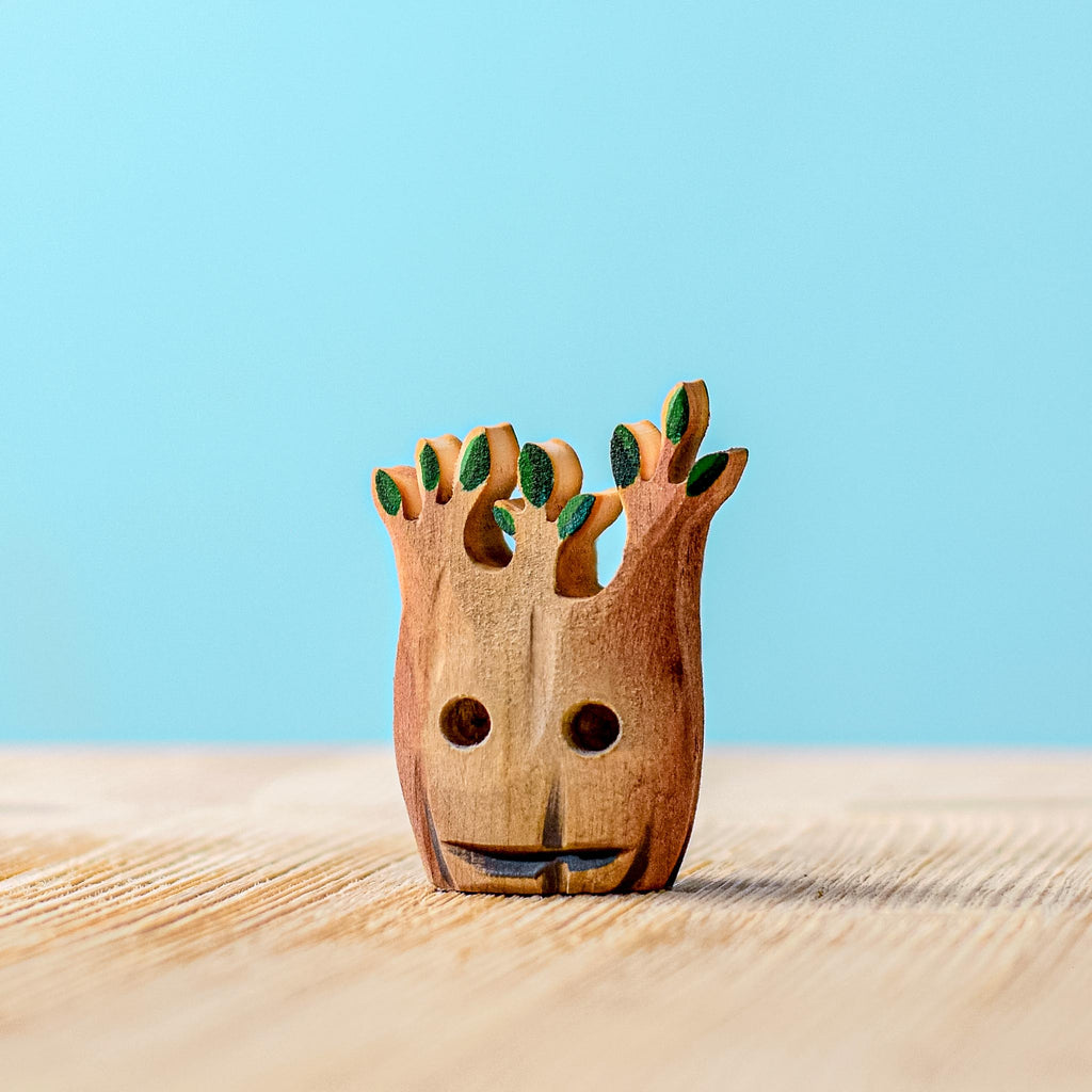 A small, handcrafted wooden figurine from the "Handmade Wooden Halloween Trees & Bat Set" collection, reminiscent of a tree with a face, complete with a smiling expression and green leaves on its branches, sits on a wooden surface against a blue background. Perfect as Halloween decor, this piece adds charm to any spooky-themed setting.