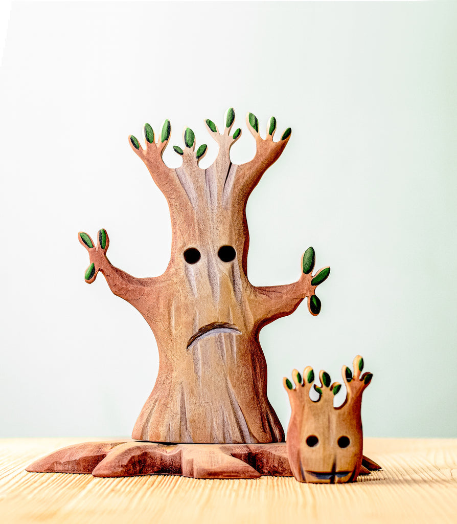 A small, handcrafted wooden sculpture from the Handmade Wooden Halloween Trees & Bat Set features two stylized trees with faces. The larger tree has a sad expression, while the smaller one in front appears content or smiling. Perfect as unique Halloween decor, they rest on a light-colored wooden surface against a plain, light background.