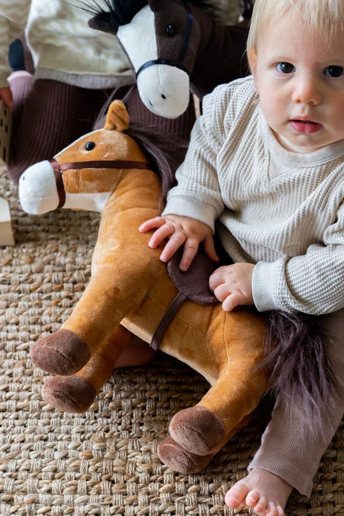 A baby sitting on a carpet with Stormy the Horse Stuffed Animal, suitable for infants.