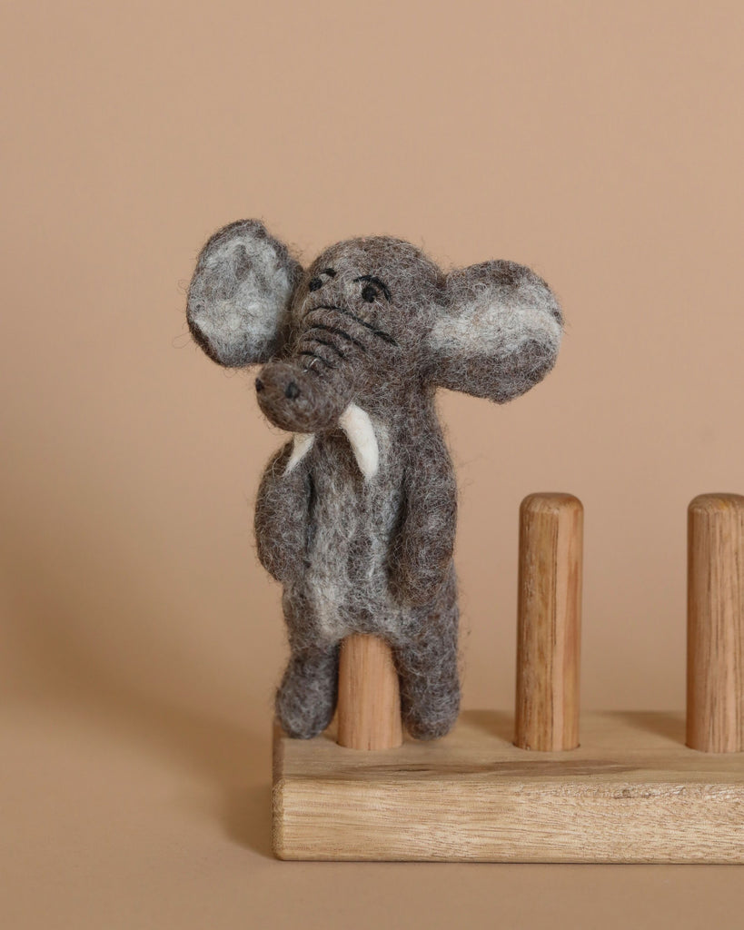 A small, gray felt "Elephant Finger Puppet" with white tusks and large ears stands on a wooden stand against a beige background. Handmade in Nepal from 100% wool, the finger puppet appears to be meticulously crafted and detailed, showcasing a slightly textured surface. The wooden stand features vertical pegs.