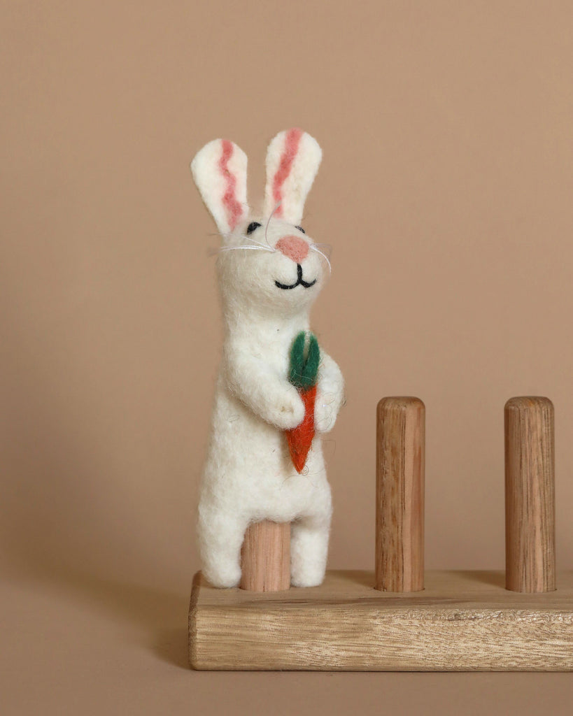 The Bunny Finger Puppet is a small, white needle-felted rabbit with pink ears that stands upright while holding a felt carrot. Handmade in Nepal, the bunny finger puppet is displayed on a wooden stand with two vertical wooden pegs, set against a plain beige background.