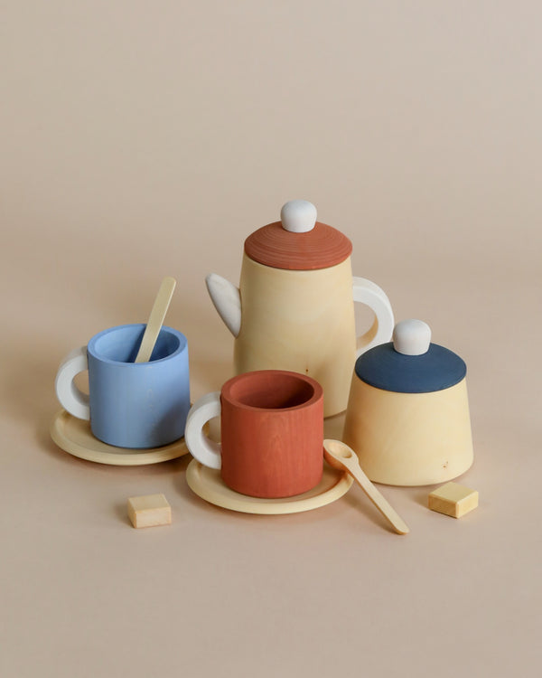 A charming Raduga Grez Handmade Wooden Tea Set, featuring a non-toxic wooden teapot with lid, and two cups – one terracotta and one blue – each with white handles and spoons. The set includes two saucers and two wooden cubes, all against a light beige background.