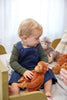 A toddler with curly blonde hair sits on a bed, dressed in a green long-sleeve shirt and blue overalls. The child clutches The Orangutans Stuffed Animal, with other plush toys, including another monkey, visible in the background. The cozy scene evokes the warmth of a mother-daughter bond.