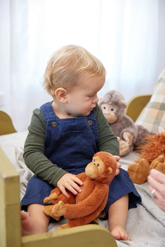 A toddler with curly blonde hair sits on a bed, dressed in a green long-sleeve shirt and blue overalls. The child clutches The Orangutans Stuffed Animal, with other plush toys, including another monkey, visible in the background. The cozy scene evokes the warmth of a mother-daughter bond.