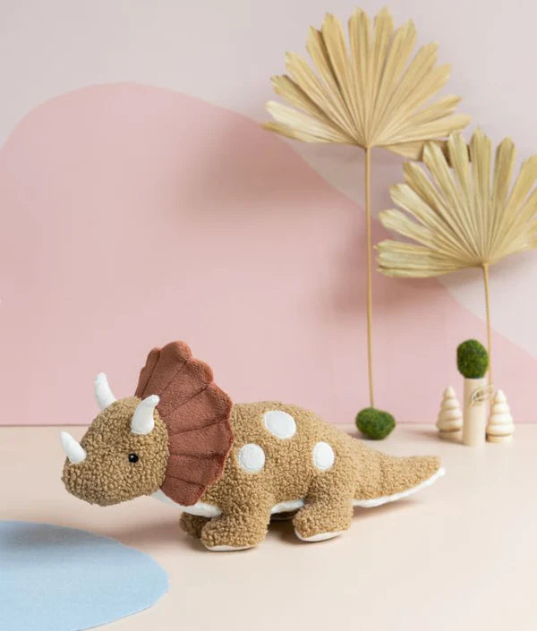 A Triceratops Dinosaur Stuffed Animal stands on a pastel surface. The 40cm x 18cm plush toy is brown with white spots and features three white horns. In the background, dried palm fronds and small green moss balls sit against a wall decorated with pink and beige geometric patterns. Suitable for ages 0+.