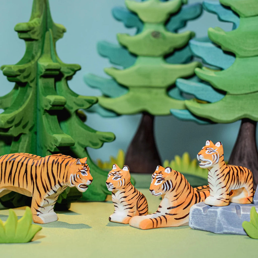 A group of four handcrafted wooden tigers from the Bumbu Wooden Tiger Set, painted in bright orange with black stripes, are placed in a forest setting. Three tigers are seated while one stands. The background features green trees and bushes, creating an imaginative play scene with the Bumbu Wooden Tiger Set.