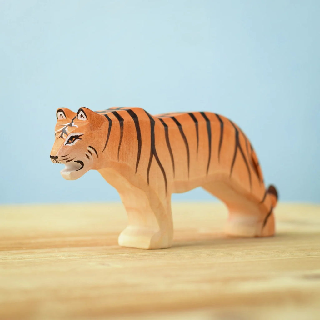 A small wooden figurine of a tiger with an orange body, black stripes, and white details on the face. Handcrafted from eco-friendly materials, this piece is part of the Bumbu Wooden Tiger Set. The figure stands on a light wooden surface against a light blue background.