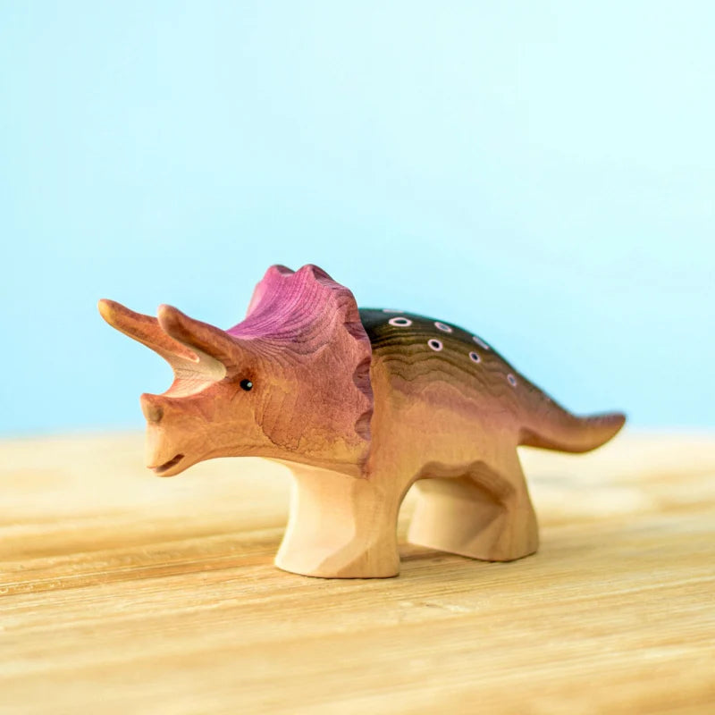 A Bumbu Handmade Triceratops Dinosaur Set crafted from sustainably sourced wood is placed on a light wooden surface with a light blue background. The toy features painted details, including a pink frill and brown and green body with white spots, all created with non-toxic paint.