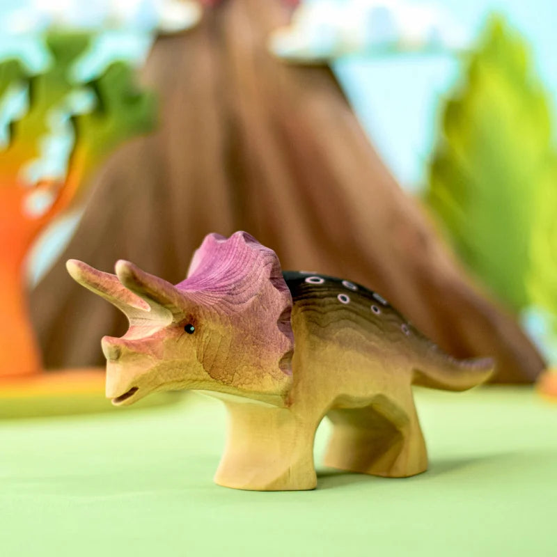 A Bumbu Handmade Triceratops Dinosaur Set, crafted from sustainably sourced wood and adorned with non-toxic paint in vibrant brown, yellow, and purple hues, stands on a green surface. Blurred, colorful trees and plants in the background suggest a playful, nature-themed setting.