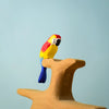 A small, brightly colored Bumbu Wooden Amazonian Parrot figurine with red, yellow, blue, and white markings perched on a smooth wooden branch against a plain light blue background.