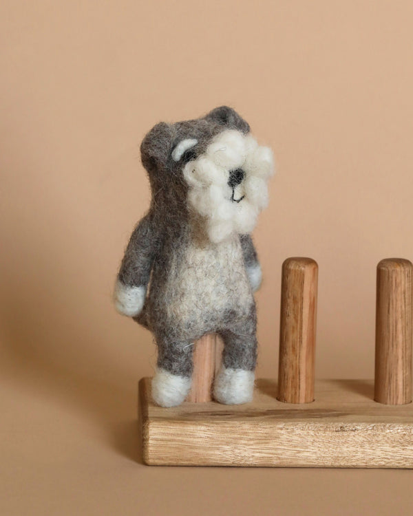 A small needle-felted Schnauzer Dog Finger Puppet, resembling a gray and white dog, stands on a wooden pegboard. The background is a plain, pale beige surface. The finger puppet features a white snout and paws, with a fluffy face and small, round black eyes.